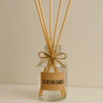 aromatizador de ambiente natural de vidro com líquido transparente e palitos difusores, decorado com laço de sisal e etiqueta com a palavra serenidade, sobre prateleira branca