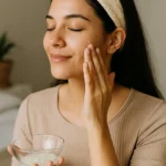 Mulher aplicando hidratação caseira no rosto com expressão relaxada e tigela de vidro com creme natural nas mãos