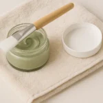 Pote de vidro com máscara facial de argila verde sobre toalha bege, com pincel de cerdas brancas e tampa ao lado